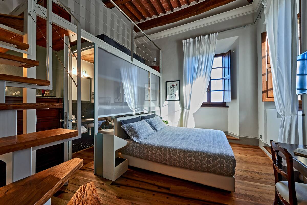 A bedroom with a bed, wooden floor, built-in shelving, a desk, and a window with curtains.