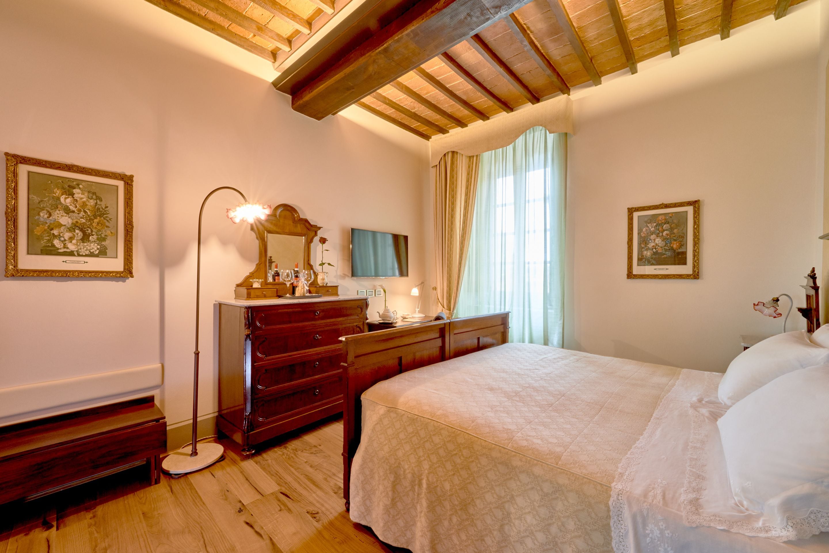 A bedroom with a wooden ceiling, a large bed, a dresser, a mirror, and framed floral artwork on the walls.