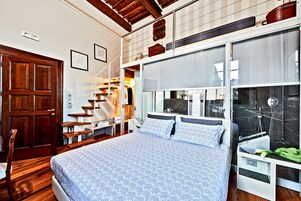A bedroom with a large bed, wooden flooring, a staircase, and a bathroom with a mirror and sink.