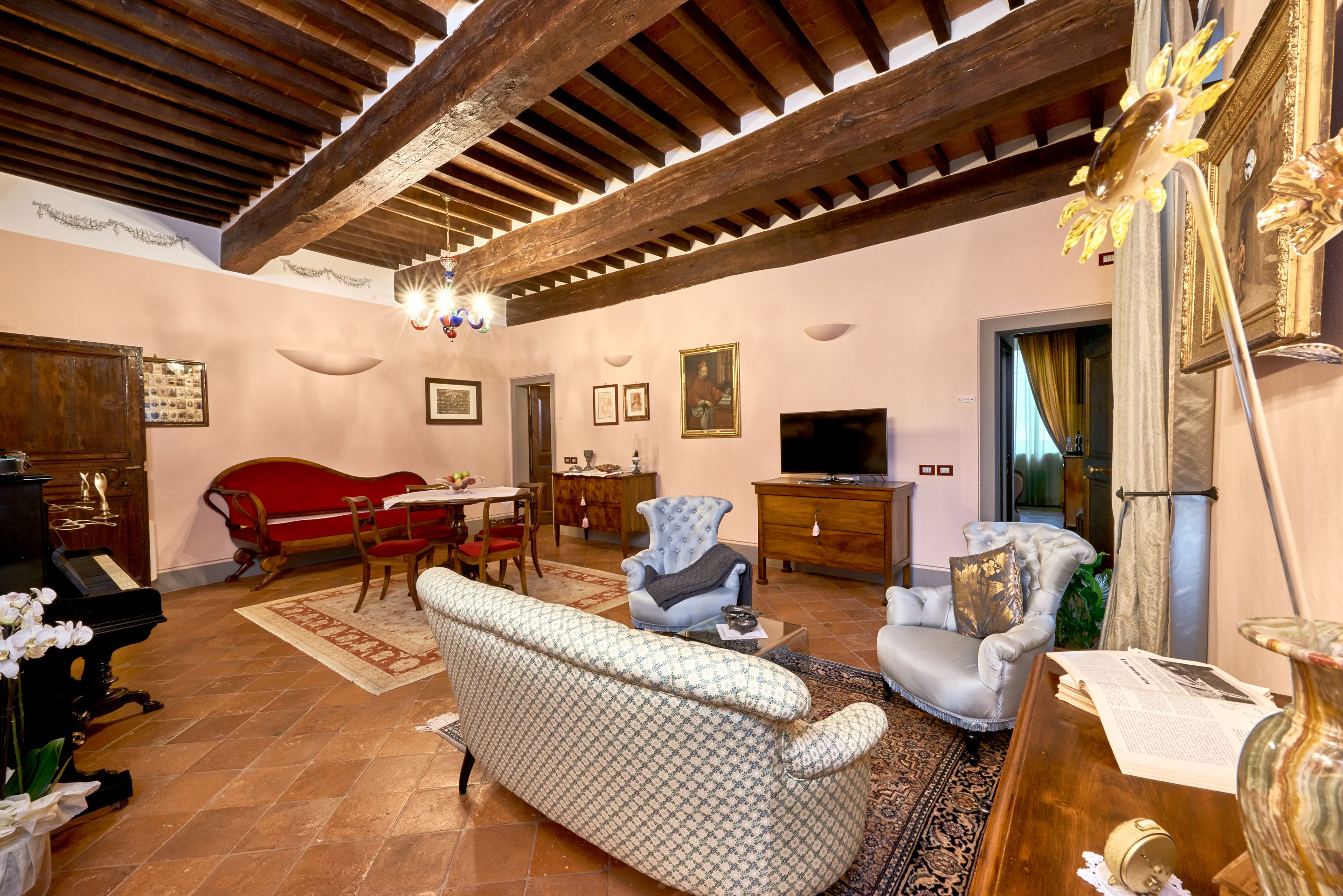 A spacious living room with a high ceiling, a grand piano, a red sofa, a patterned sofa, a television, and a dining area with a table and chairs.