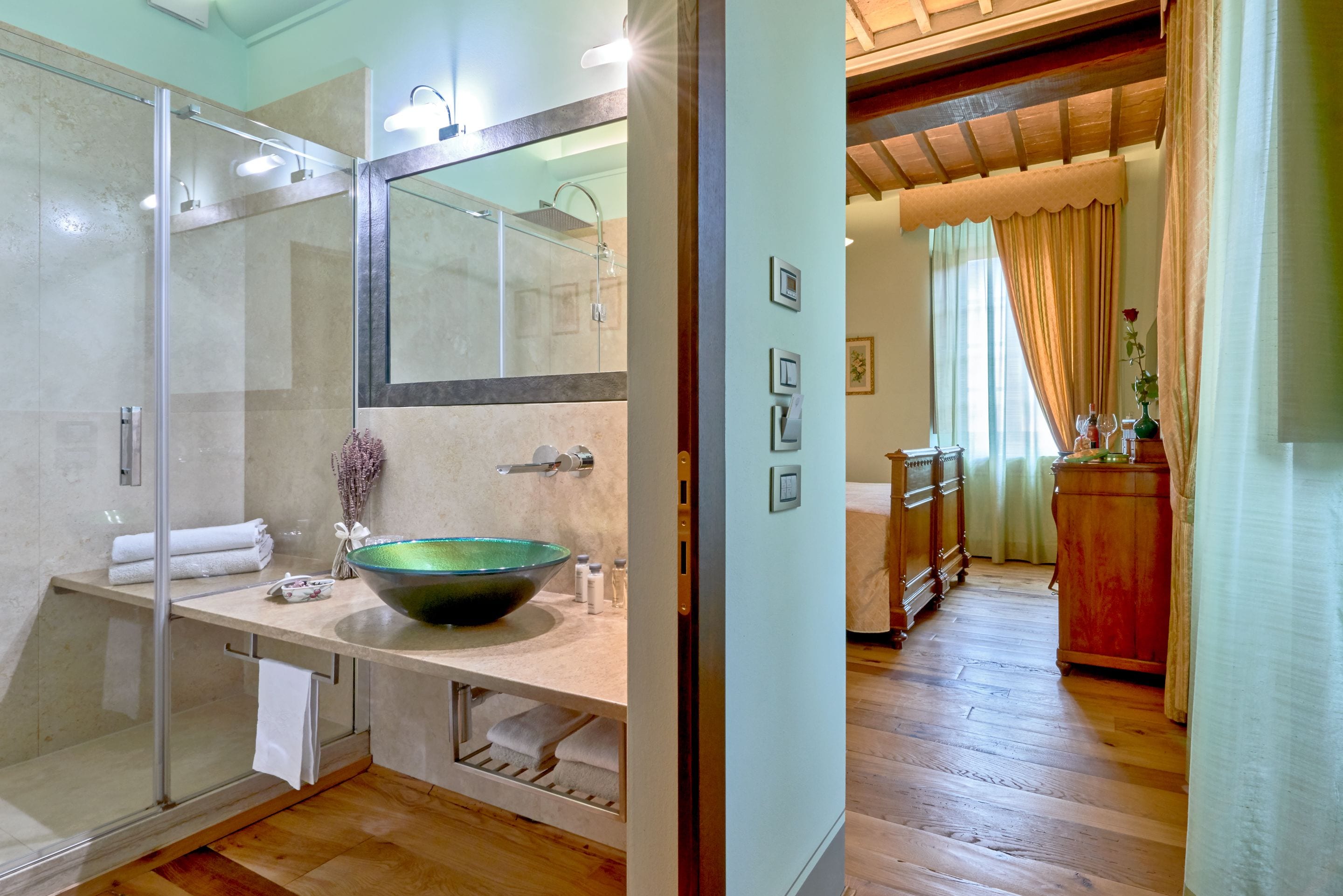 A modern bathroom with a glass-enclosed shower, a large mirror, a green bowl sink, and wooden flooring.
