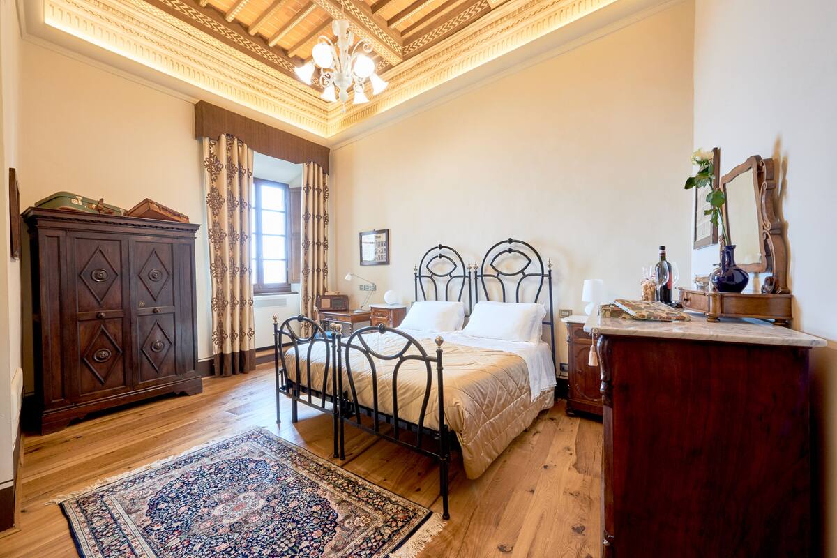 A bedroom with a wooden floor, a bed with a headboard, a nightstand, a dresser, a mirror, and a window with curtains.