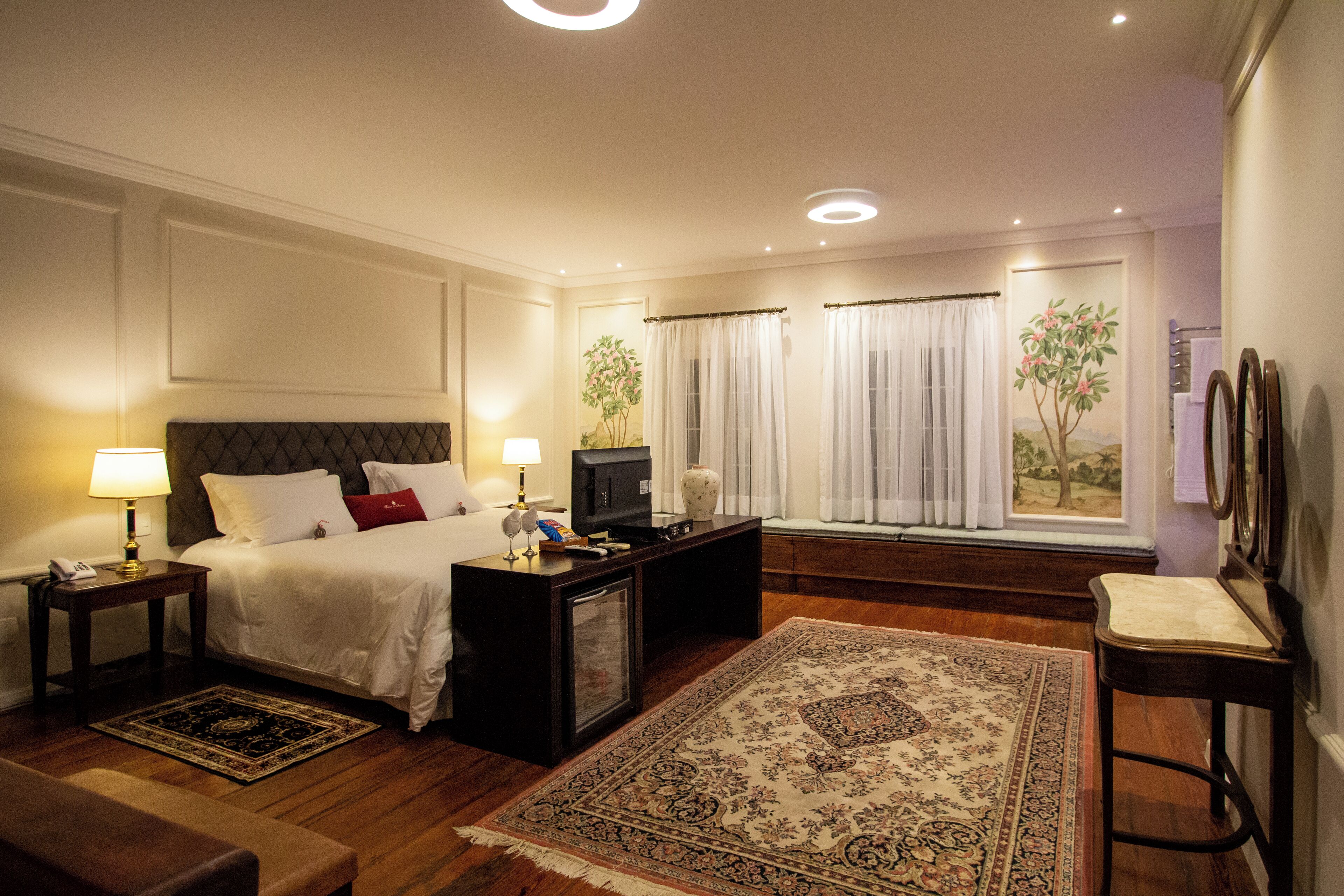 A hotel room with a bed, a television, a desk, a chair, a rug, and a window with curtains.