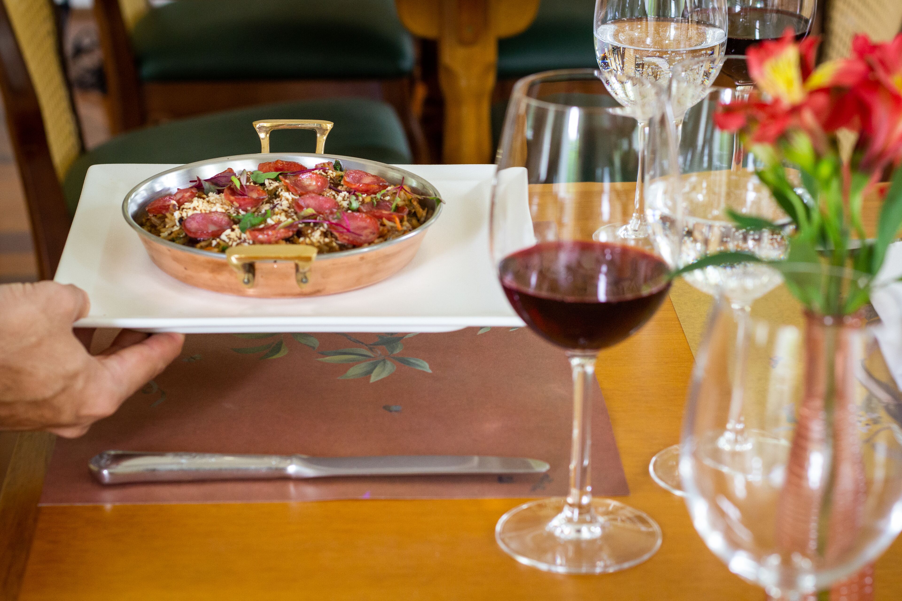 A dining table with a metal pan of food, a wine glass, and a vase with flowers.