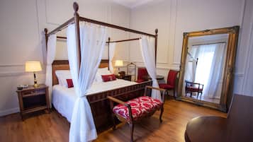 Een bed met hemelbed, voorzien van witte lakens, een rode bekleden bankje, een nachtkastje met lamp en een grote spiegel aan de muur.