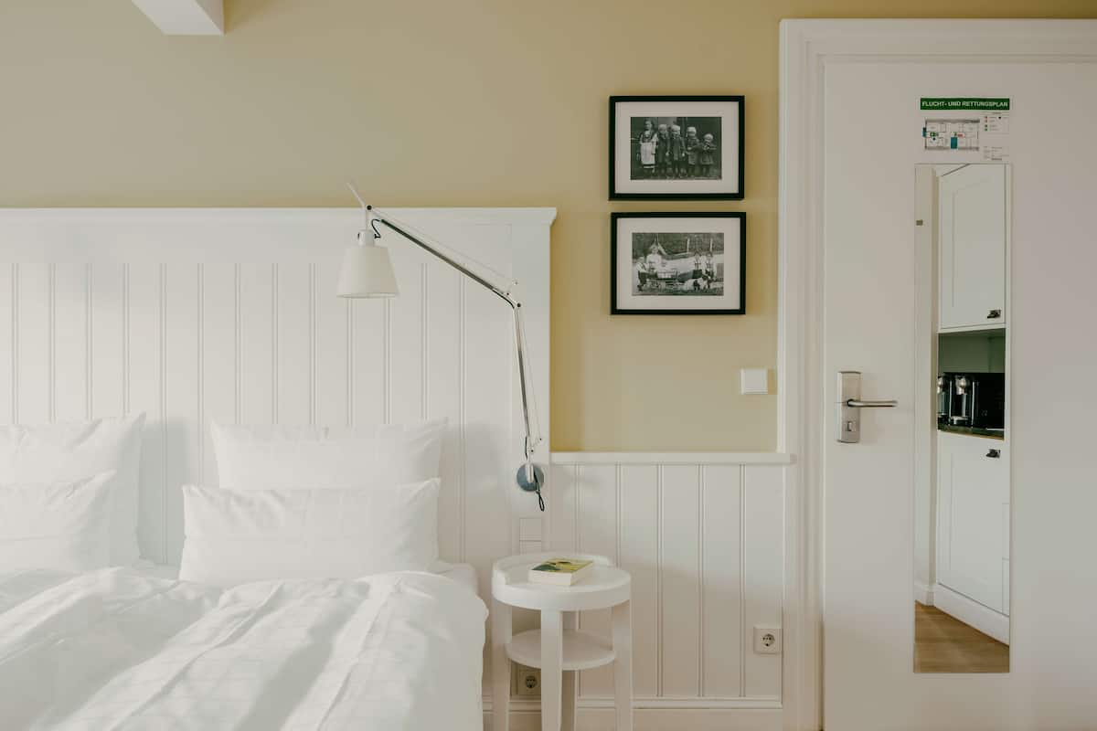 A bedroom with a white bed, a bedside lamp, a small round table, and two framed photographs on the wall.