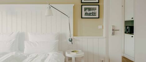 A bedroom with a white bed, a bedside lamp, a small round table, and two framed photographs on the wall.