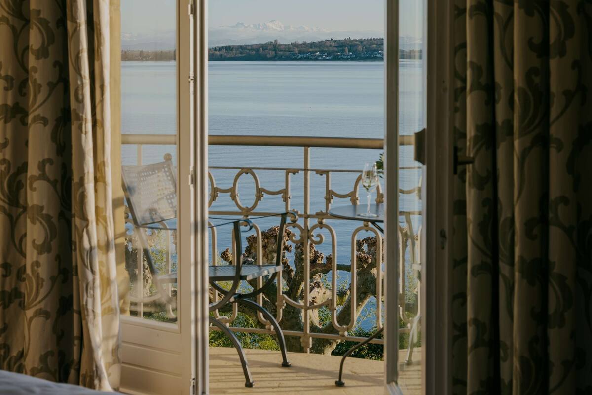 A room with a bed and a balcony with a view of a lake and mountains.