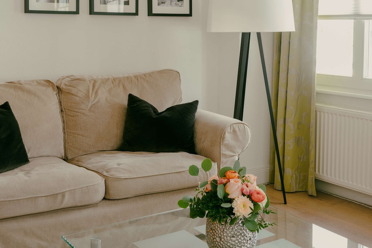 A modern living room with a beige sofa, black cushions, a glass coffee table, a vase with flowers, and three framed photographs on the wall.