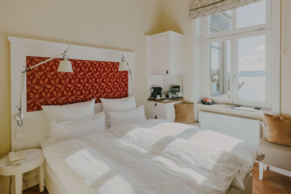 A neatly made bed in a room with a large window, a desk with a computer, and a telephone on a side table.