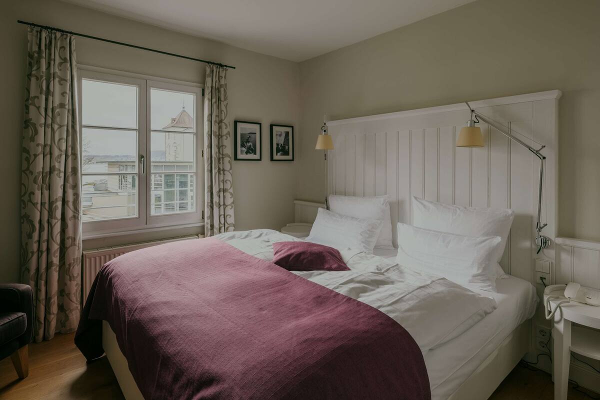 A bedroom with a large bed, a bedside table with a lamp, a window with curtains, and a view of a building outside.