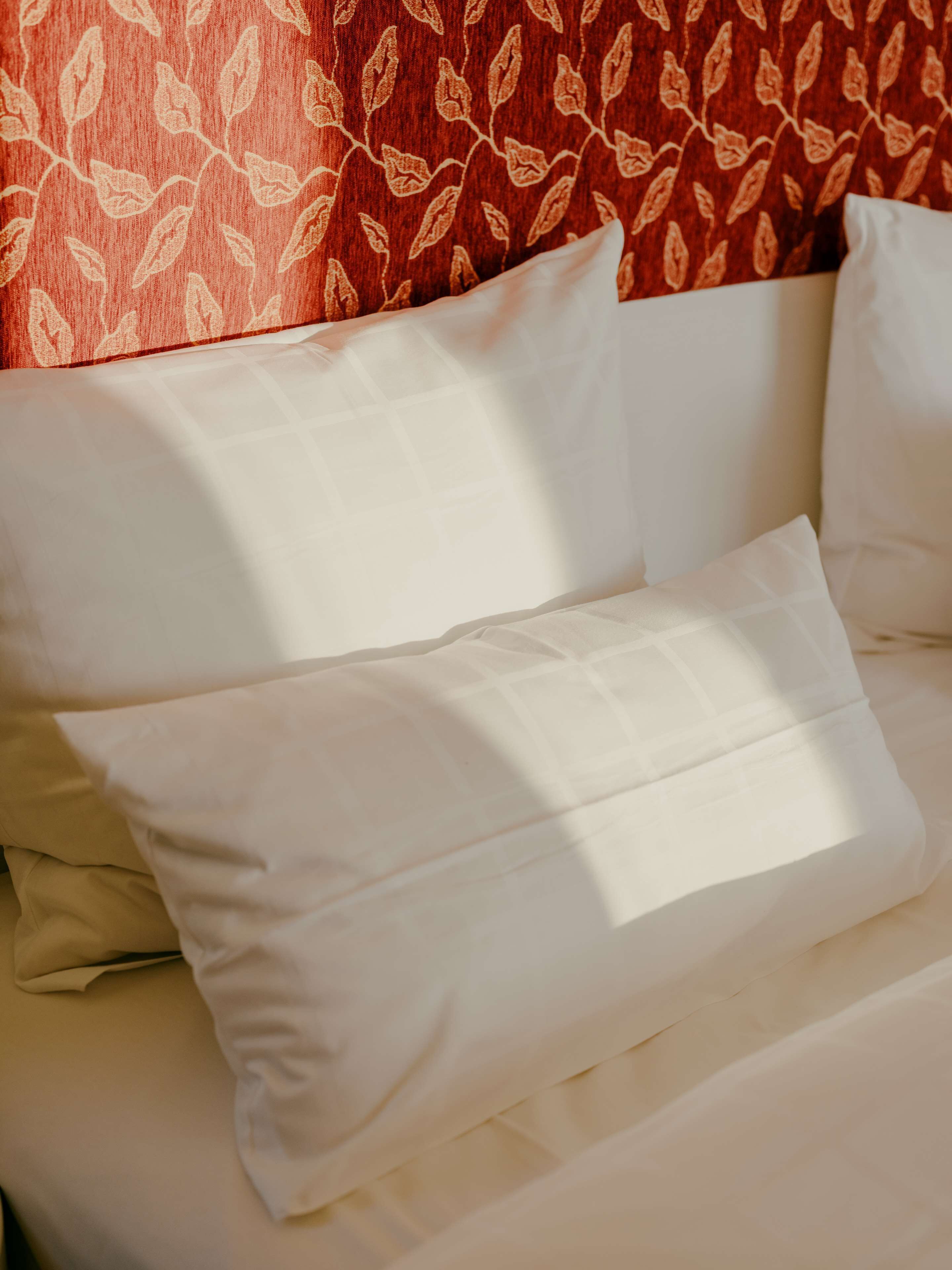 Una cama con ropa blanca y una pared con estampado rojo detrás.