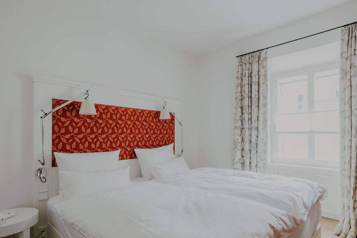 A neatly made bed with white linens, a red patterned headboard, and a bedside table with a lamp.