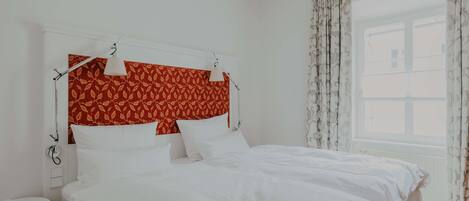 A neatly made bed with white linens, a red patterned headboard, and a bedside table with a lamp.