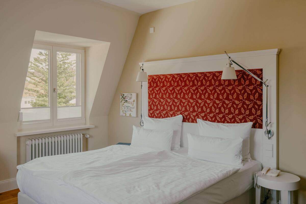 A bedroom with a bed, a window, a radiator, a bedside table with a lamp, and a telephone.
