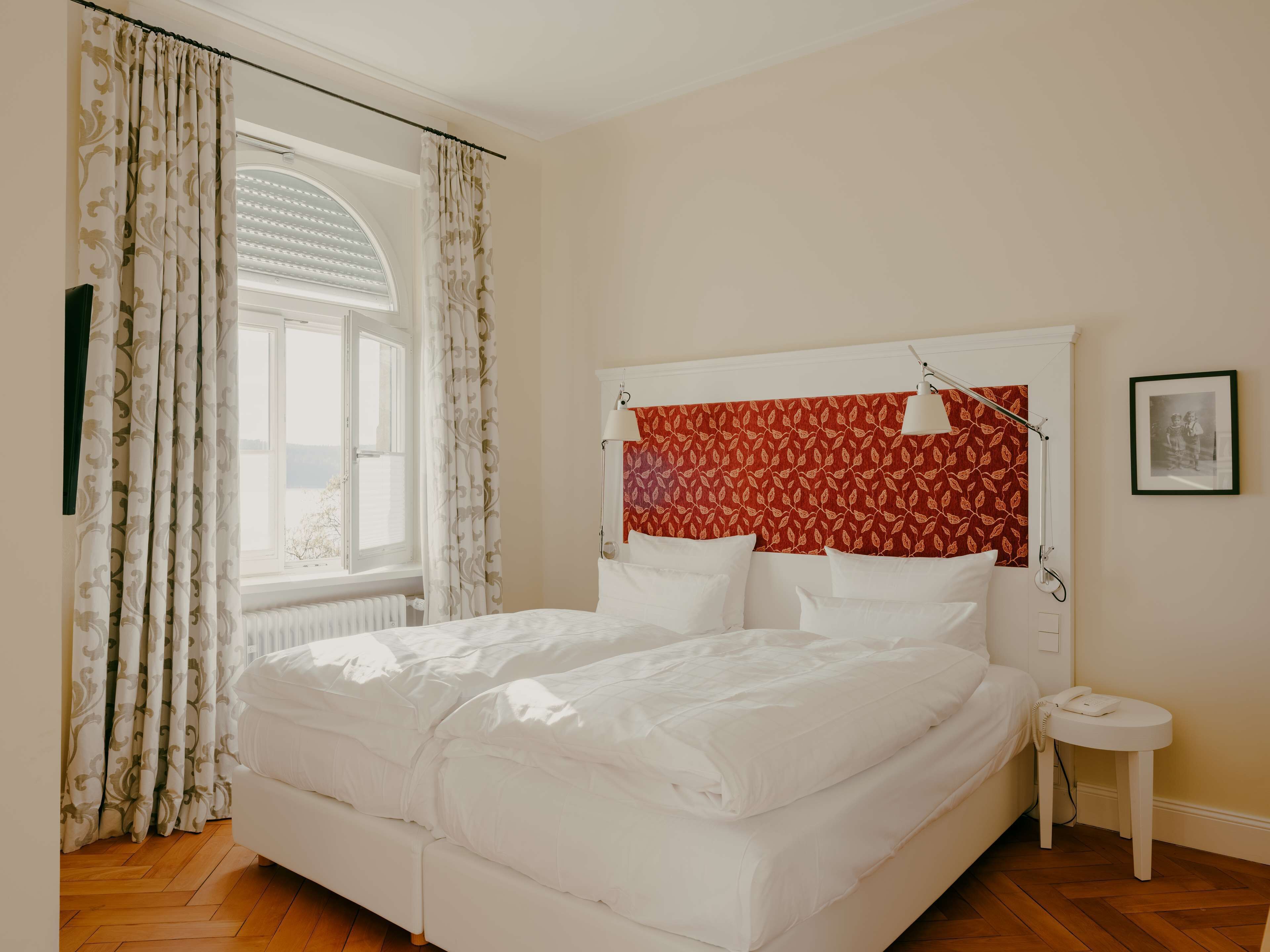 A neatly made bed with white bedding, a red patterned headboard, and a small bedside table with a lamp and phone.