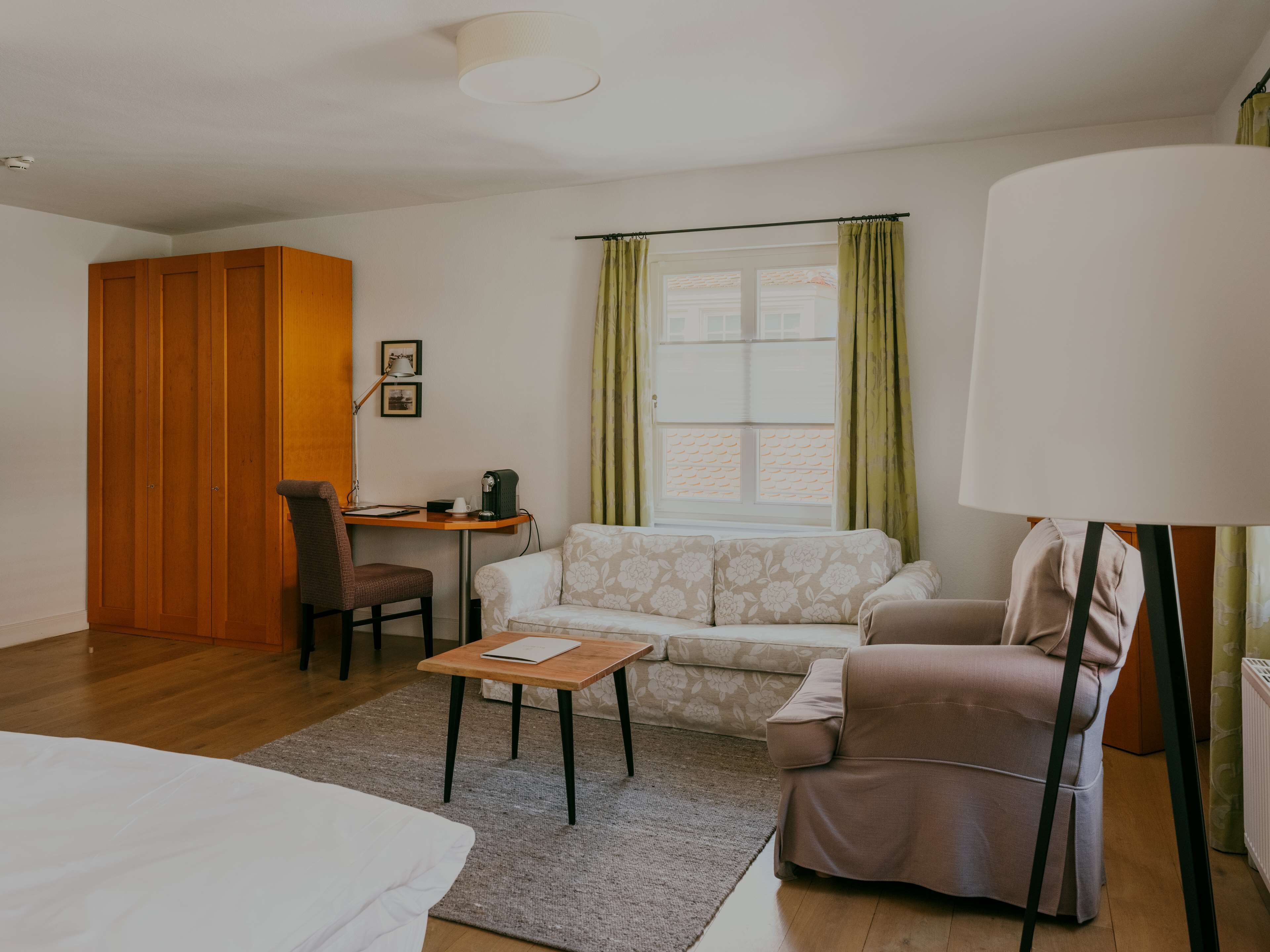 Ein kleines Hotelzimmer mit Bett, Schlafsofa, Sessel, Tisch, Schreibtisch und einem Fenster mit Vorhängen.