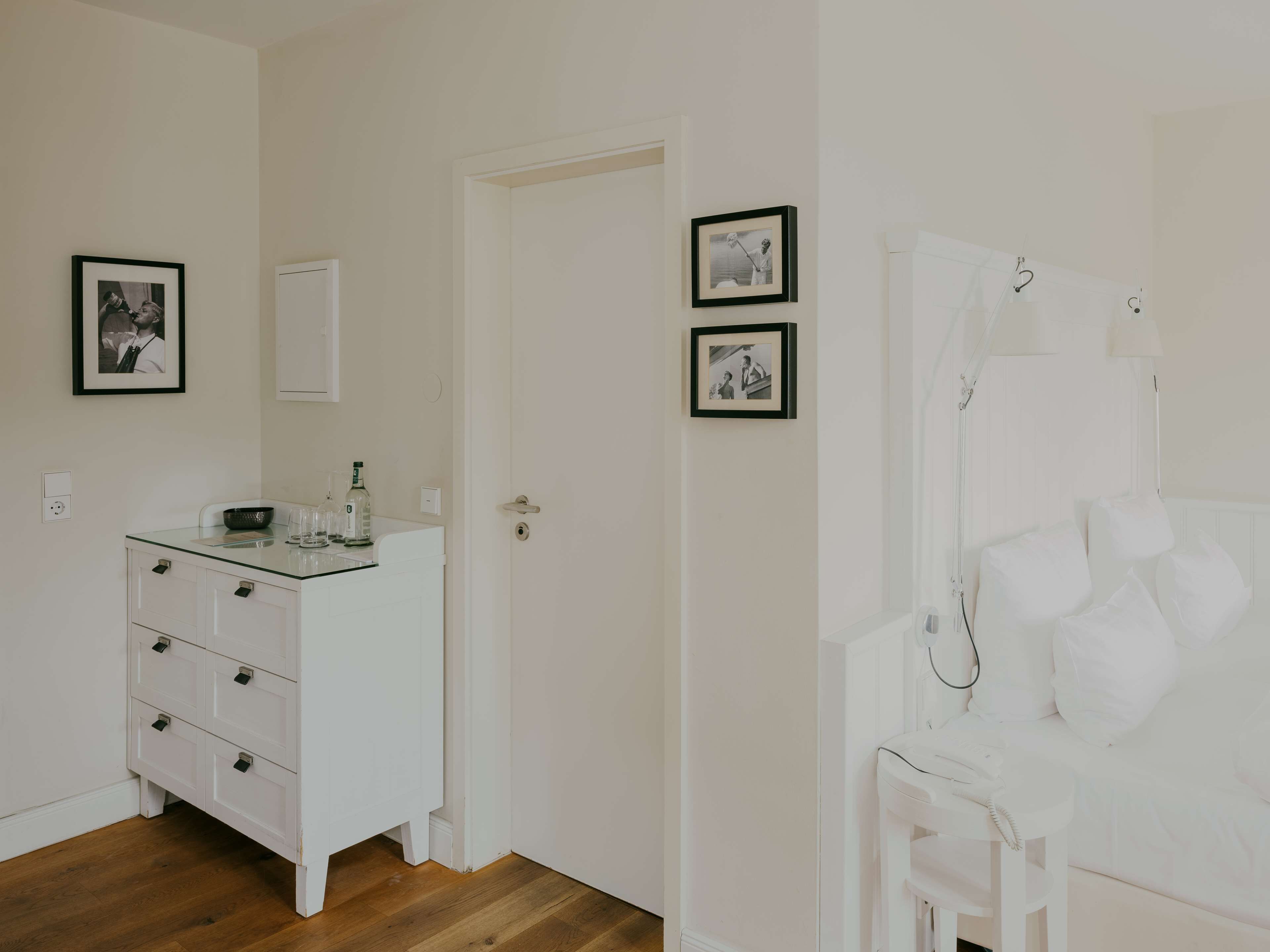 Una habitación con un aparador blanco, una silla blanca y una cama con ropa de cama blanca. Hay dos cuadros en la pared.