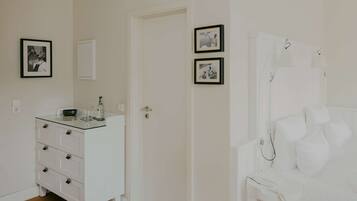 Una habitación con un aparador blanco, una silla blanca y una cama con ropa de cama blanca. Hay dos cuadros en la pared.