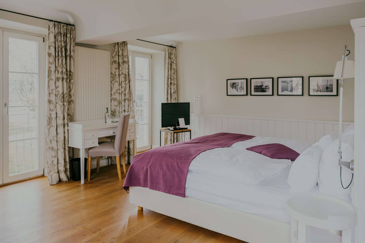 A bedroom with a bed, a desk, a chair, a TV, and a window with curtains.
