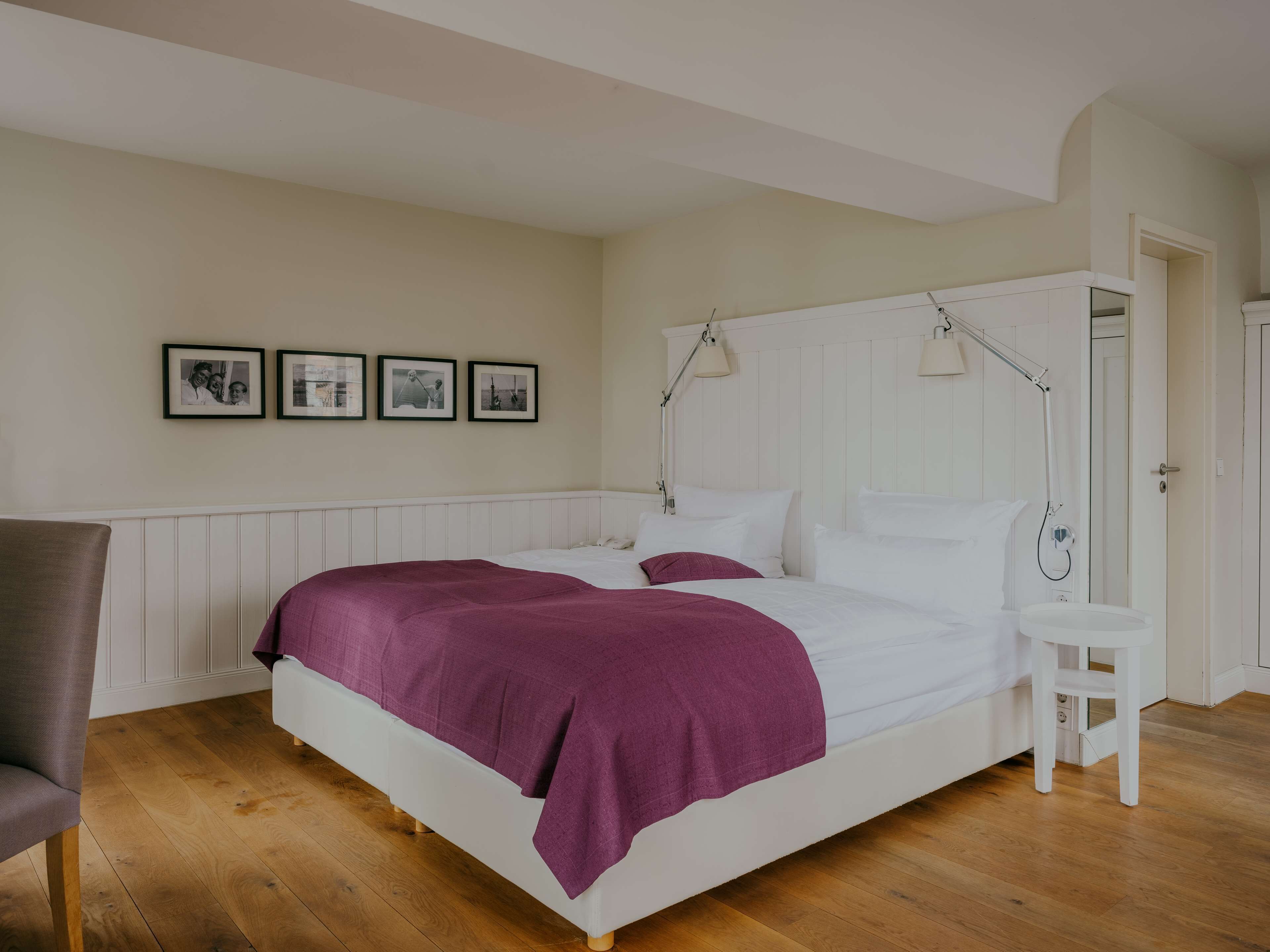 A bedroom with a large bed, a bedside table, a chair, and a door. There are three framed pictures on the wall.