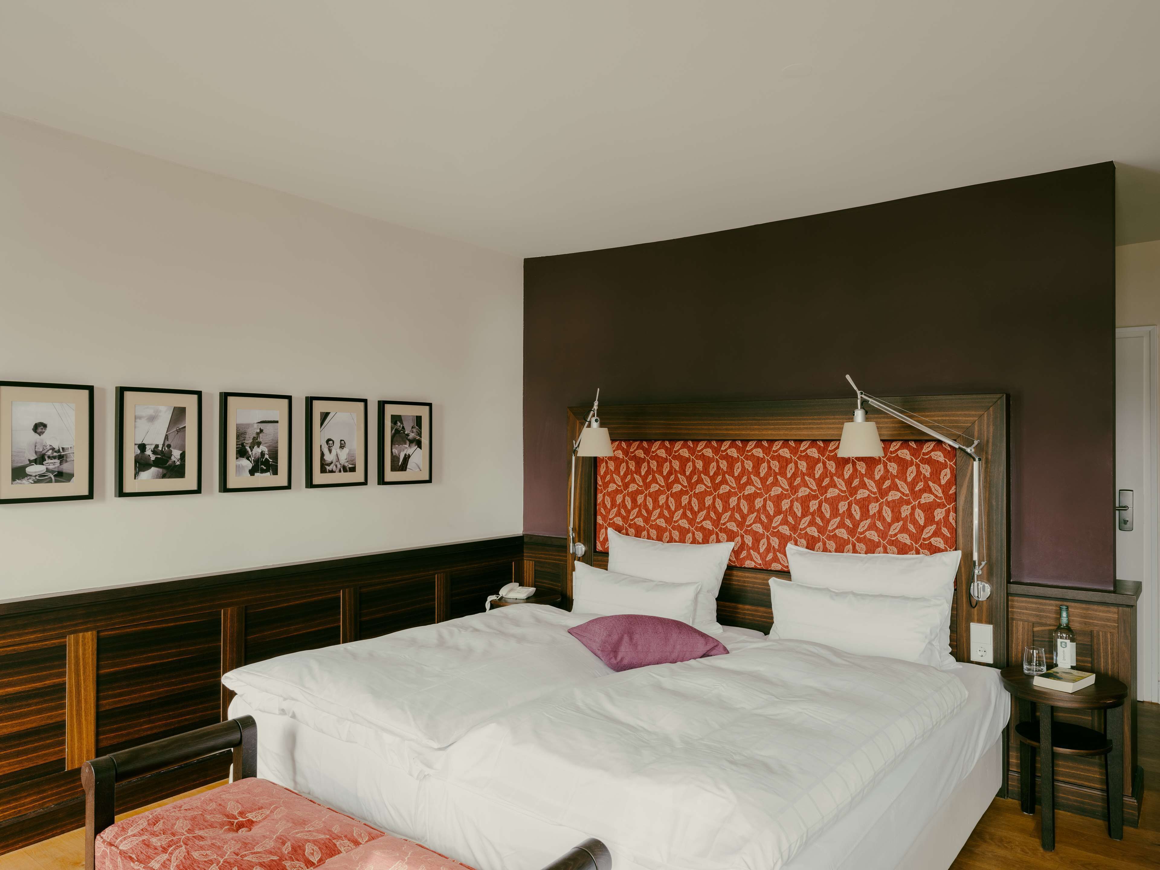 Habitación de hotel con una cama grande, una mesita de noche con lámpara, una pequeña mesa con una botella y vasos, y cuatro cuadros enmarcados en la pared.