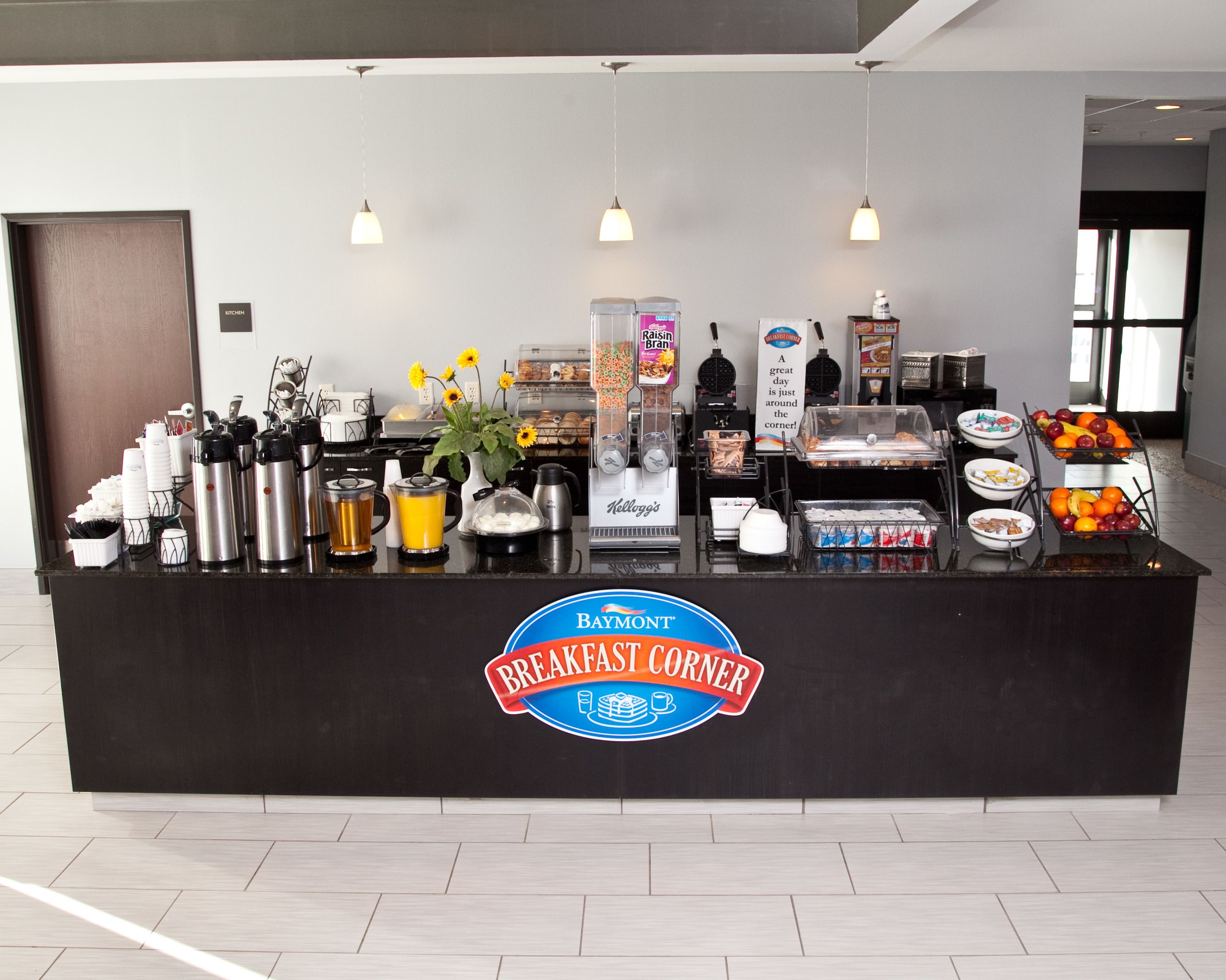 A breakfast counter with coffee machines, a fruit display, and a sign for Baymont Breakfast Corner.