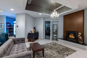 Un salon moderne avec un canapé gris, une petite table en bois et un tapis à motifs. On y trouve une cheminée ornée d’une statue, ainsi qu’un téléviseur posé sur un meuble.
