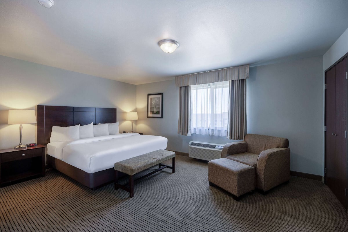 Ein Hotelzimmer mit einem großen Bett, einem Nachttisch mit Lampe, einem Sessel, einer Bank und einem Fenster mit Vorhängen.