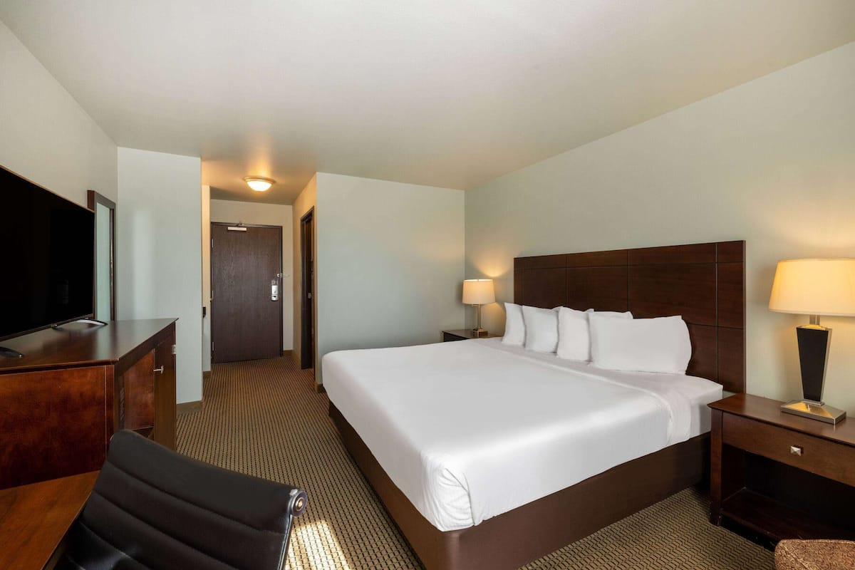 Ein Hotelzimmer mit einem großen Bett, einem Schreibtisch mit Fernseher, einem Sessel, einem Nachttisch mit Lampe und einer Tür zu einem weiteren Raum.