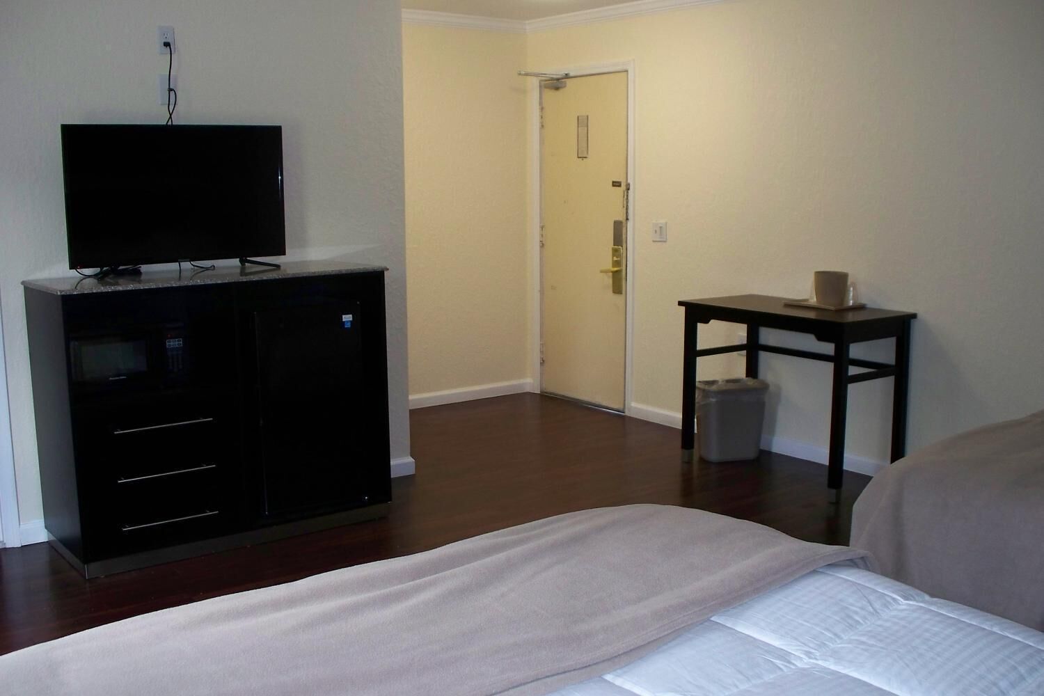 A hotel room with a bed, a TV on a cabinet, a small table with a cup, and a door in the background.