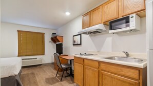 A compact kitchen with wooden cabinets, a microwave, a sink, and a dining area with a chair and table. - WoodSpring Suites College Station (Bryan)