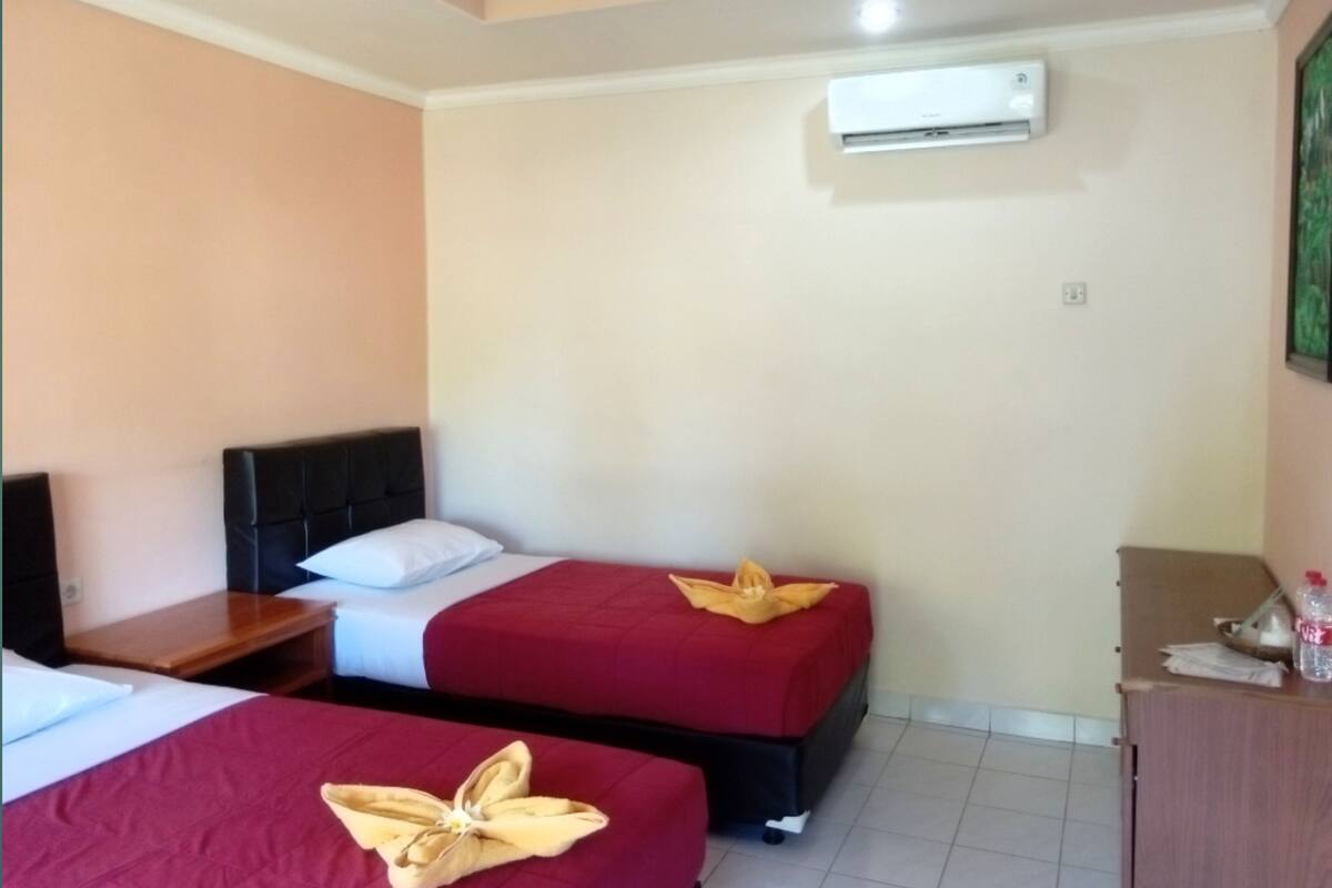 A hotel room with two beds, each with a red bedspread and a golden decorative pillow. There is a wooden nightstand with a lamp, a small wooden table, and a cabinet with a bottle on top. An air conditioning unit is mounted on the wall above the beds.