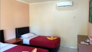 Habitación de hotel con dos camas, cada una con una colcha roja y un cojín decorativo dorado. Hay una mesita de noche de madera con una lámpara, una pequeña mesa de madera y un mueble con una botella encima. Un equipo de aire acondicionado está montado en la pared sobre las camas.