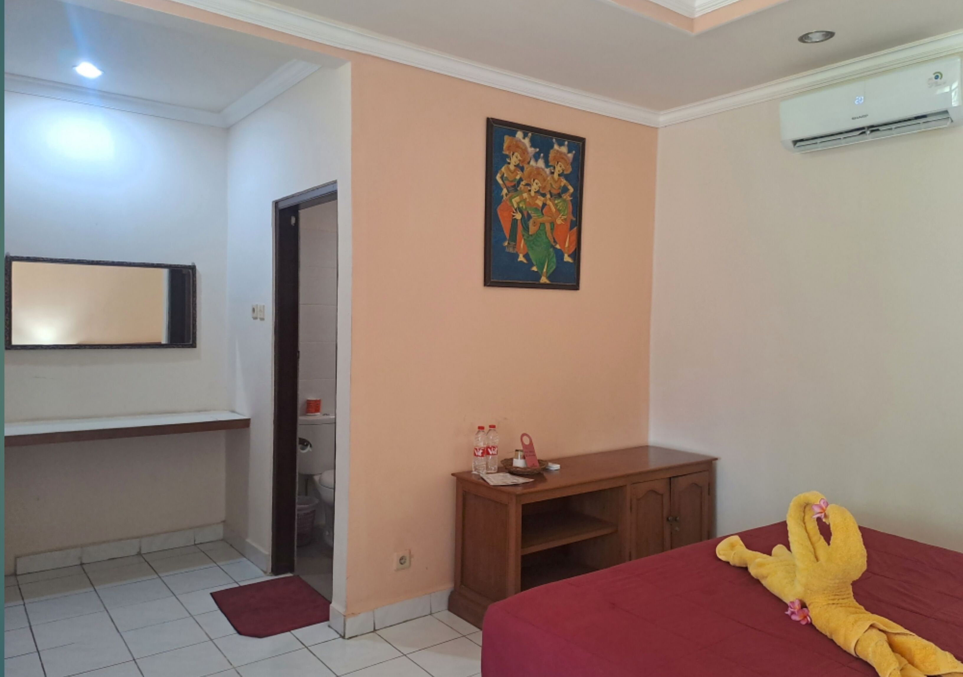 A hotel room with a bed, a desk, a chair, a painting on the wall, and an air conditioning unit.