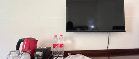 Habitación de hotel con una televisión de pantalla plana montada en la pared, una mesita con una taza roja, dos botellas de agua, un control remoto, una revista y una caja de pañuelos.