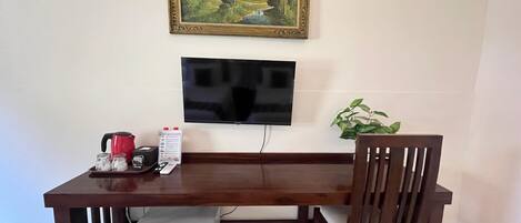 A dining area with a wooden table, a chair, a small refrigerator, a television mounted on the wall, and a painting above the TV.