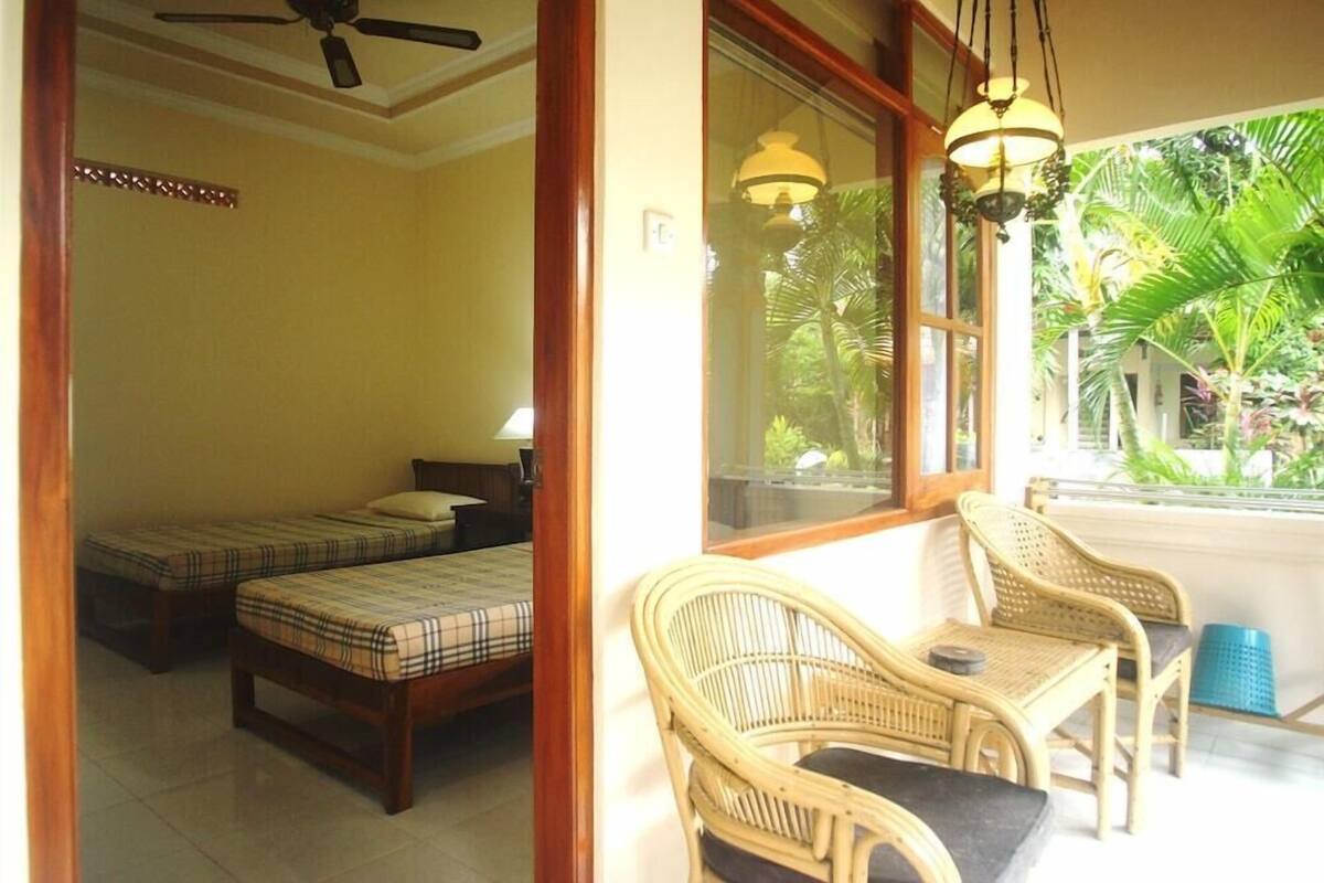 A room with two beds, a ceiling fan, and a balcony with wicker furniture.