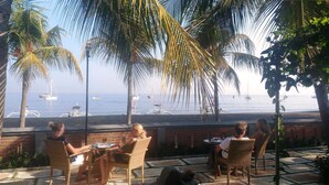 Un patio con sillas y mesas de mimbre, con vista a una playa y barcos.