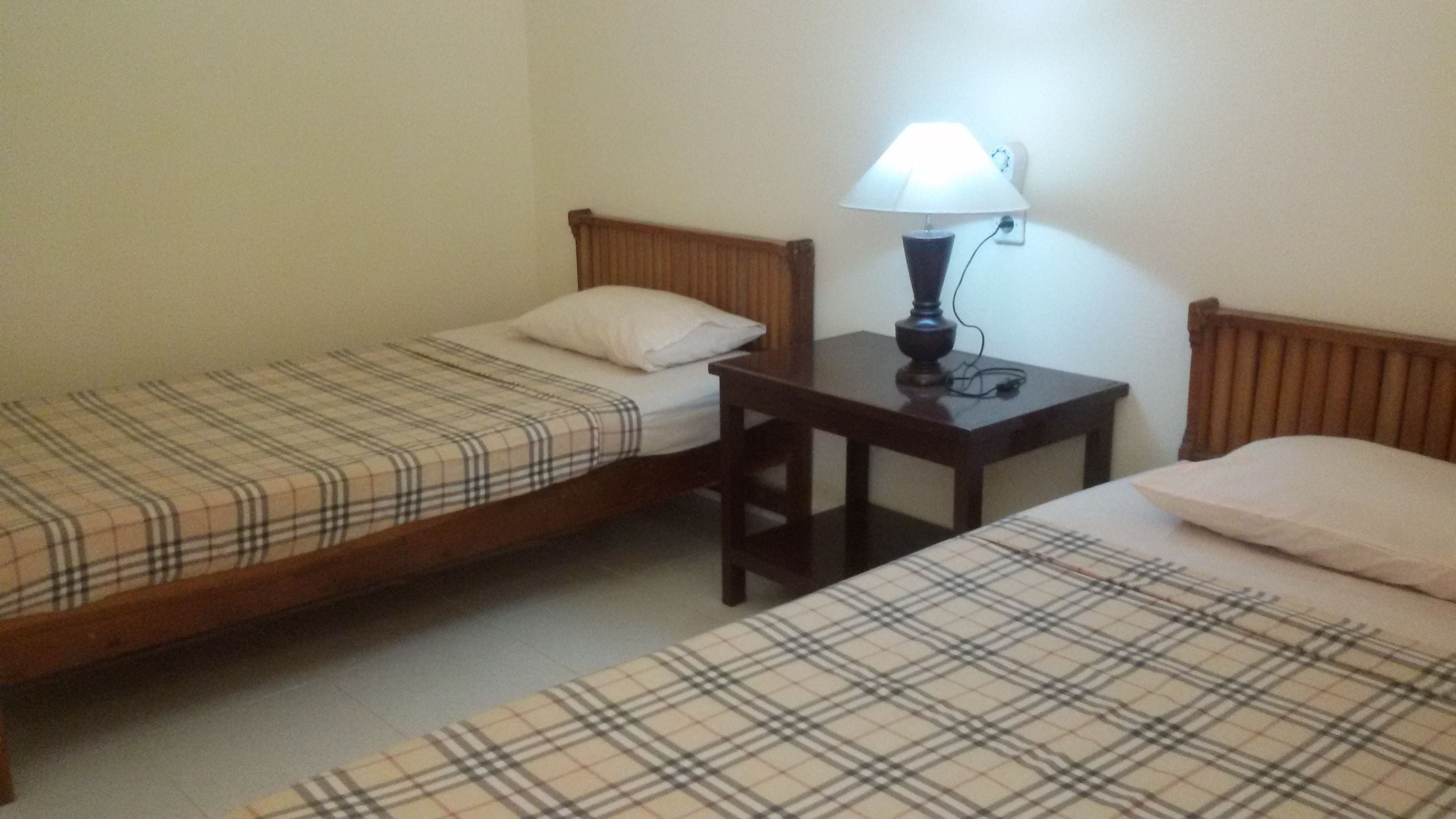 Une chambre avec deux lits simples, chacun recouvert d’une couverture à carreaux et doté d’une tête de lit en bois. Une petite table avec une lampe se trouve entre les deux lits.