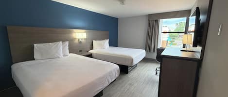 Ein Hotelzimmer mit zwei Betten, einem Schreibtisch, einem Fernseher und einem fensterbepflanzten Fenster.