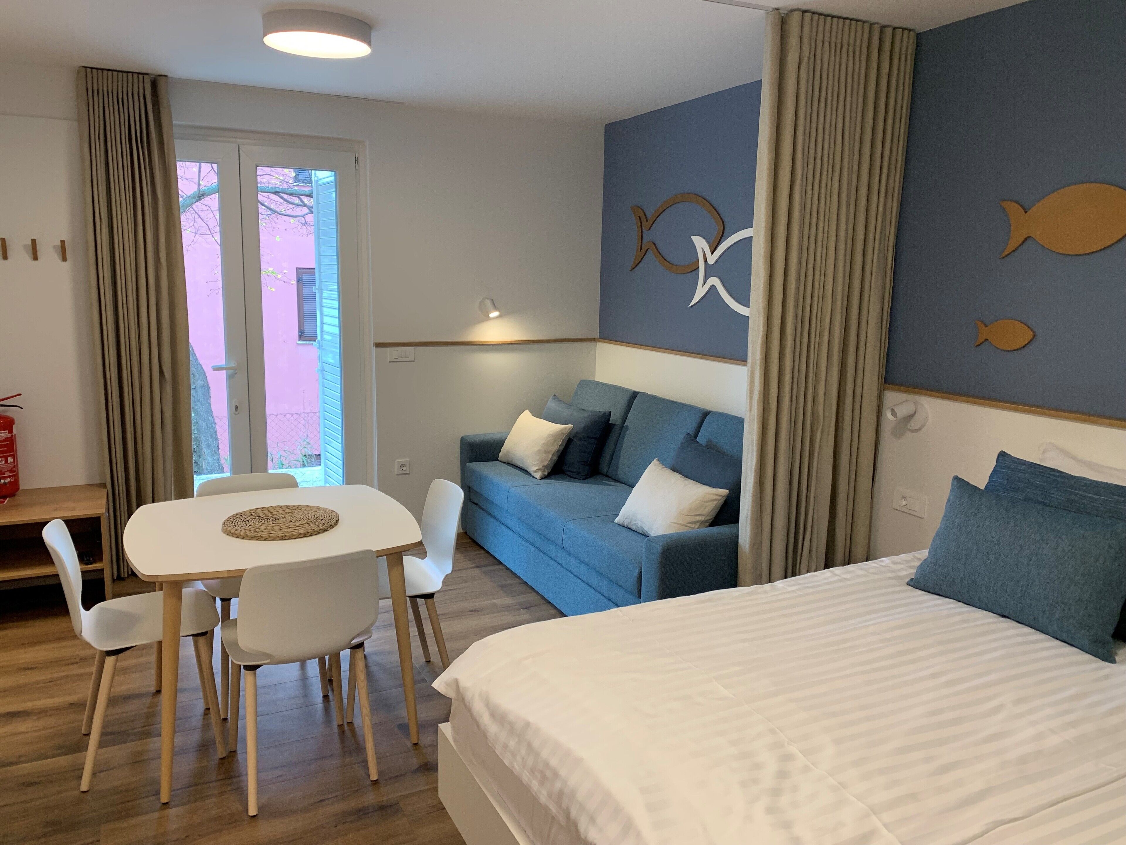 A hotel room with a bed, a blue sofa, a dining table with chairs, and a view through a door.