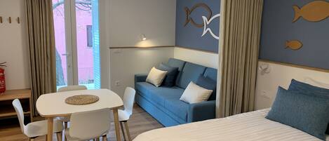 A hotel room with a bed, a blue sofa, a dining table with chairs, and a view through a door.