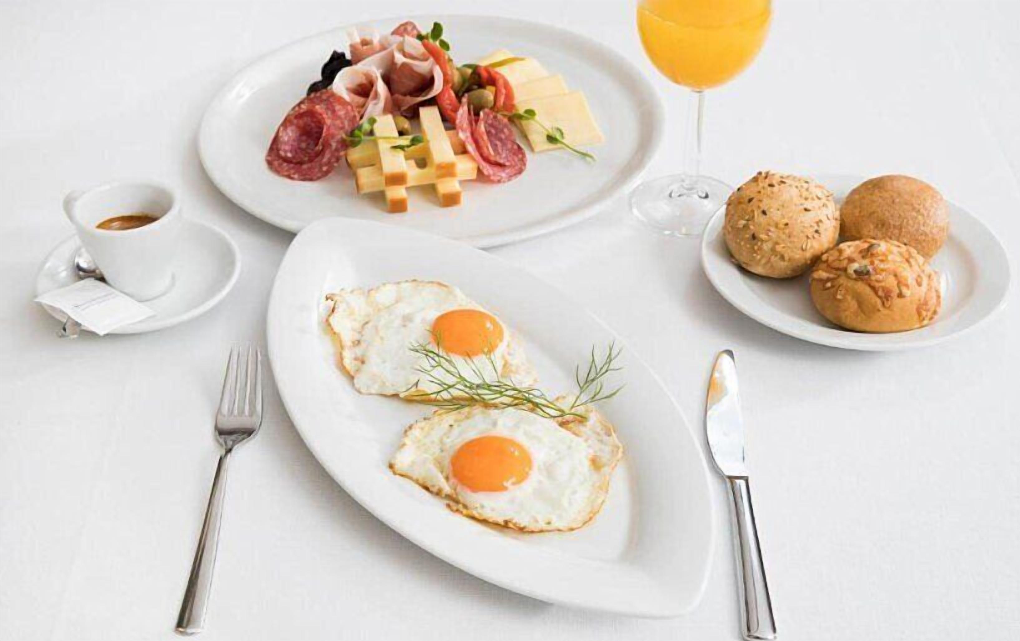 A breakfast setting with two fried eggs, a plate of meats and cheese, a glass of orange juice, and two croissants on a white table.
