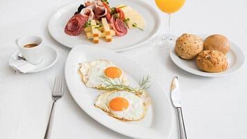 A breakfast setting with two fried eggs, a plate of meats and cheese, a glass of orange juice, and two croissants on a white table.