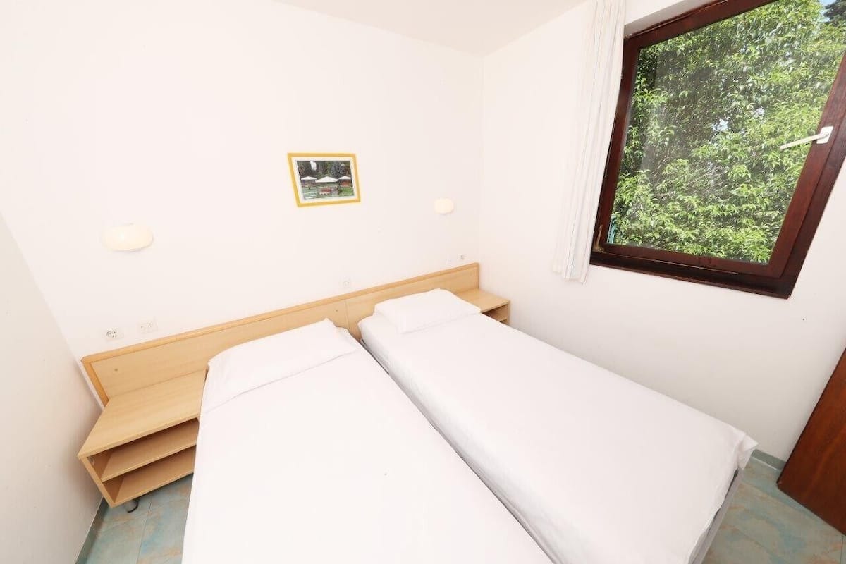 Una habitación pequeña y limpia con una cama individual, una ventana con vistas a árboles y un cuadro en la pared.