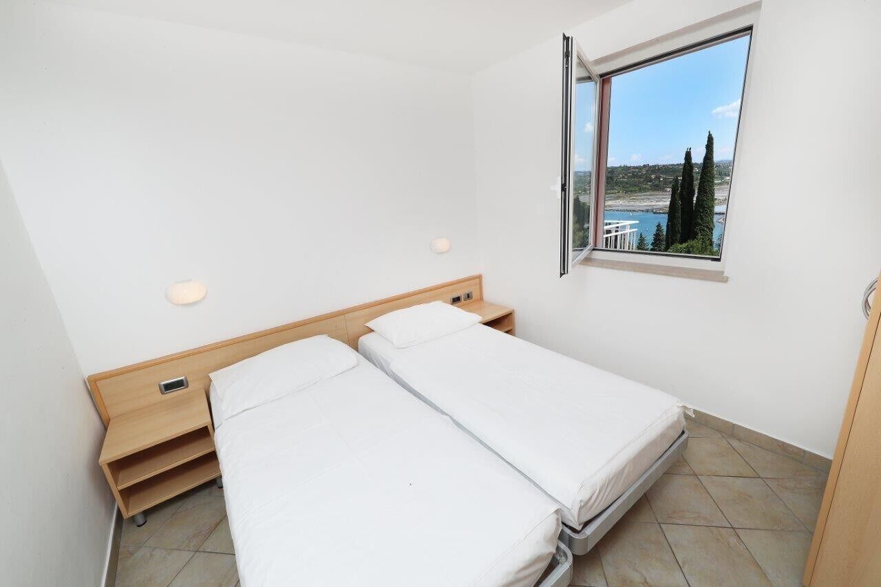 A small, clean room with two single beds, a wooden nightstand, a small table, a window with a view of trees and a body of water, and a tiled floor.
