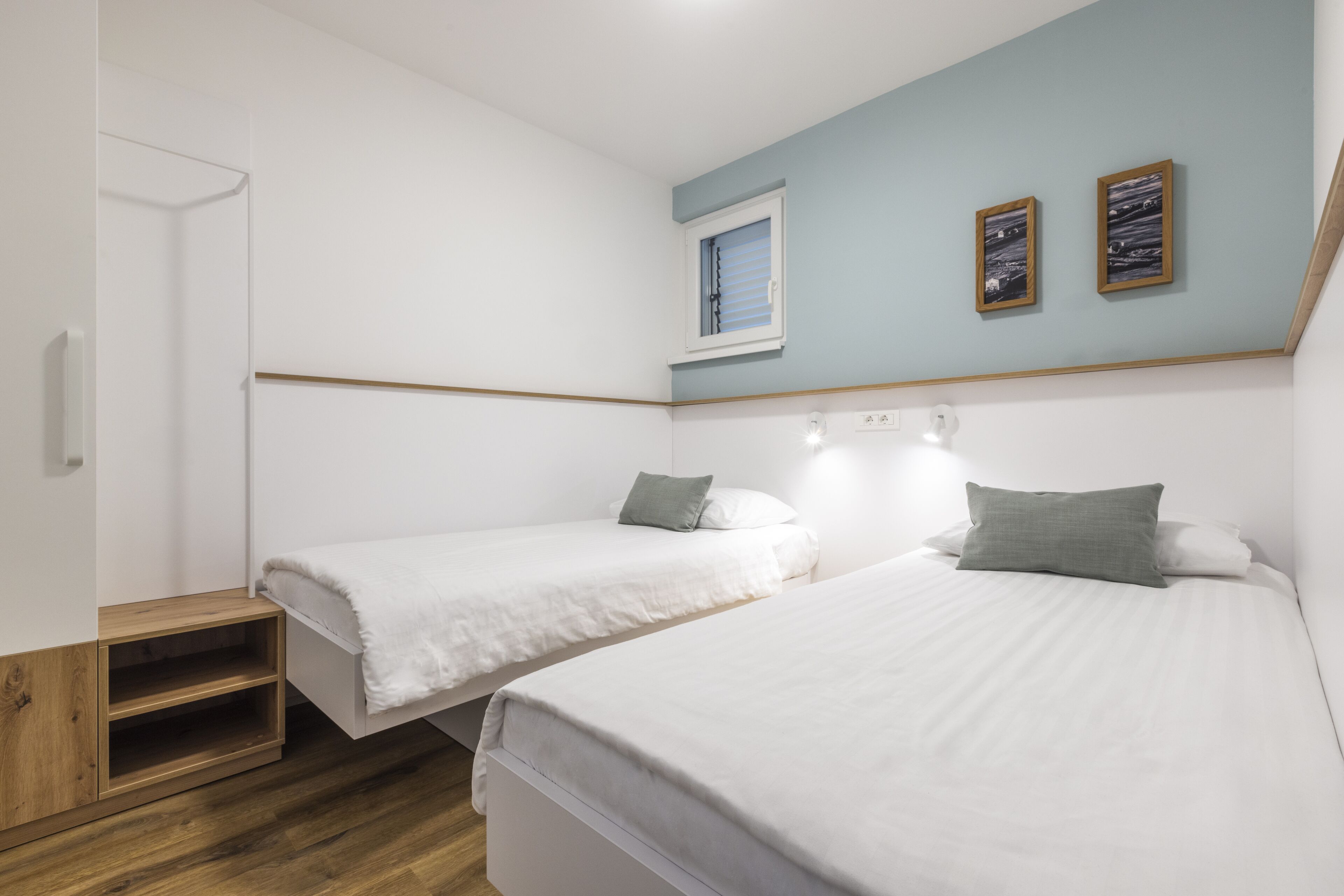 Une chambre avec deux lits, une étagère en bois, une fenêtre et deux tableaux encadrés accrochés au mur.