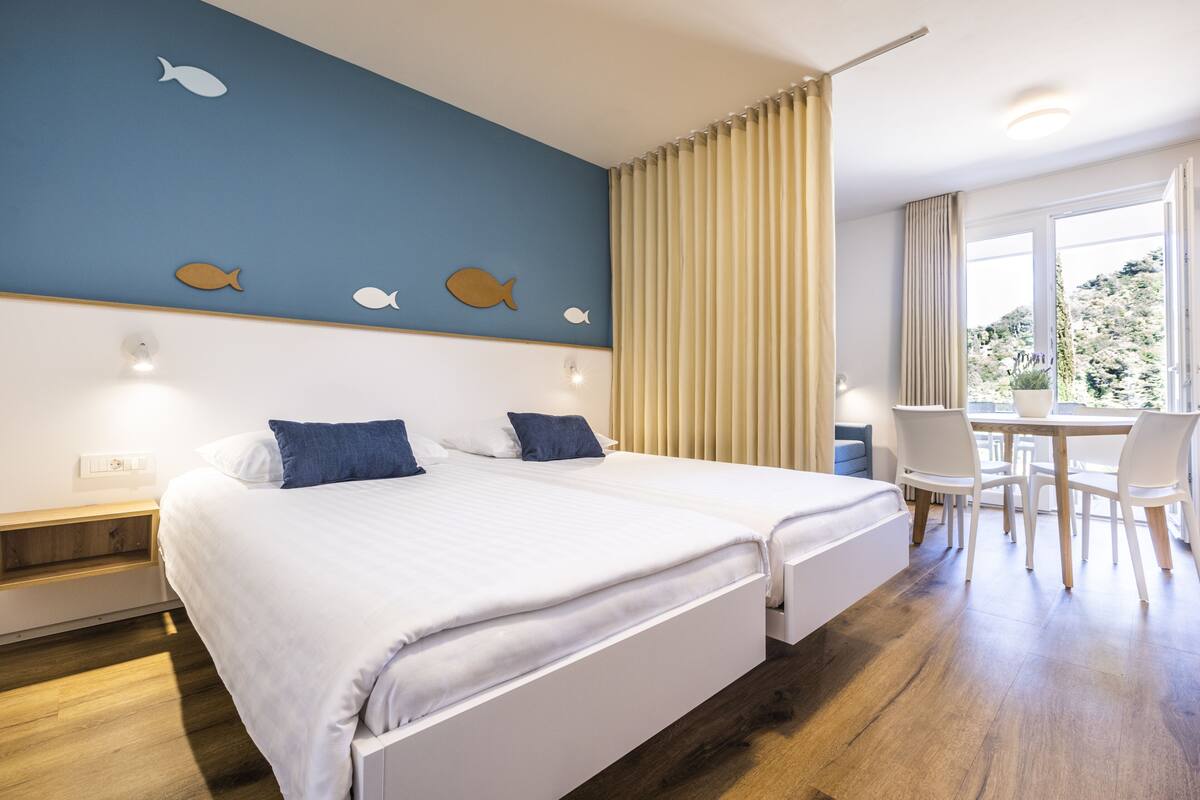 Habitación de hotel con dos camas, zona de comedor con sillas, ventana con cortinas y pared decorada con motivos de peces.