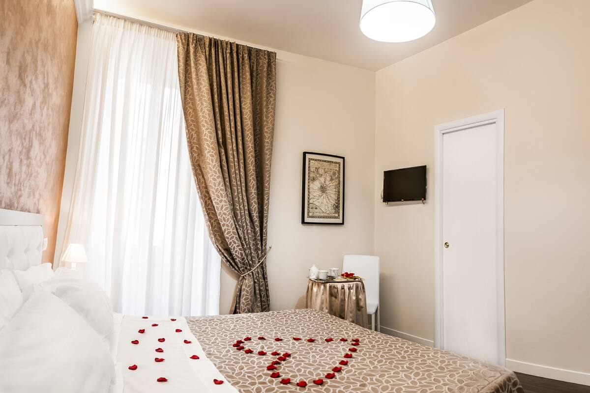 A hotel room with a bed, a bedside table with a lamp, a small table with a teapot and cups, a framed picture on the wall, and a door.